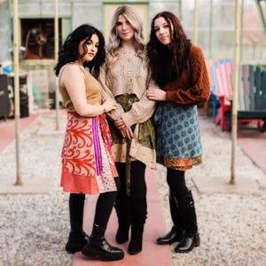 May include: Three people wearing layered outfits, including skirts and sweaters, pose outdoors. The skirts feature colorful patterns and textures. The outfits are paired with black leggings and boots. The background includes a greenhouse and colorful chairs.