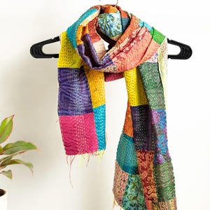 Könnte beinhalten: Ein bunter Patchwork-Schal mit einer Vielzahl von Stoffen und Mustern. Der Schal besteht aus Stoffquadraten in Gelb-, Rosa-, Blau-, Lila- und Grüntönen. Er hängt an einem schwarzen Kleiderbügel.