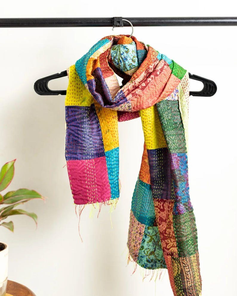 May include: A colorful patchwork scarf with a variety of fabrics and patterns. The scarf is made of squares of fabric in shades of yellow, pink, blue, purple, and green. It is hanging on a black hanger.