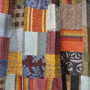 May include: A patchwork textile featuring a variety of fabrics in a range of colors including gold, orange, blue, and burgundy. The design incorporates geometric shapes and patterns, with visible stitching throughout. The textile appears to be a quilt or decorative fabric.