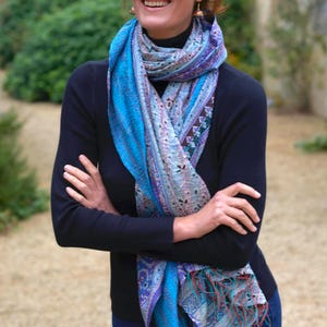 May include: A woman wearing a blue and purple patterned scarf with fringe. The scarf is draped over a black long-sleeve shirt and blue jeans. The woman is smiling with her arms crossed.
