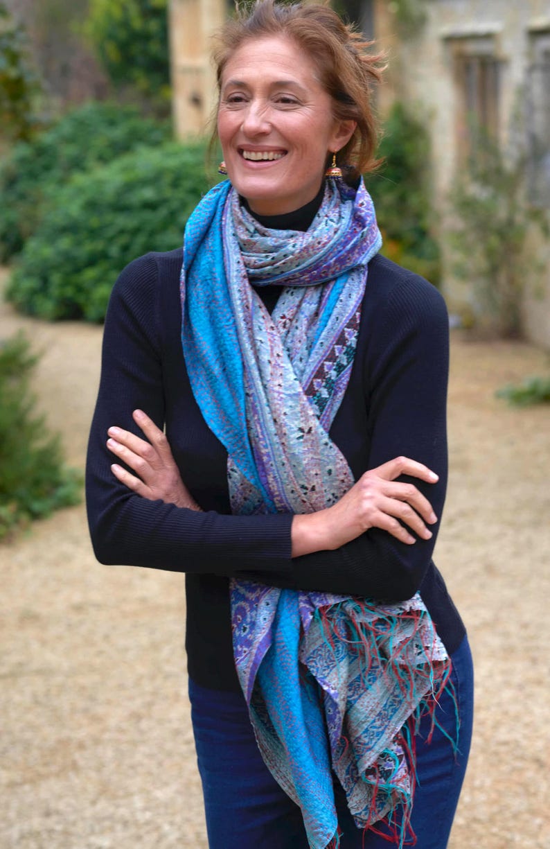 May include: A woman wearing a blue and purple patterned scarf with fringe. The scarf is draped over a black long-sleeve shirt and blue jeans. The woman is smiling with her arms crossed.