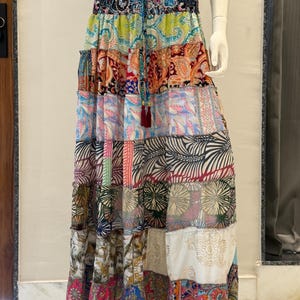 May include: A floor-length tiered skirt featuring a patchwork design with various colorful patterns including paisley, floral, and leaf motifs. The skirt has an elasticized waistband with a drawstring tie finished with two dark red tassels.