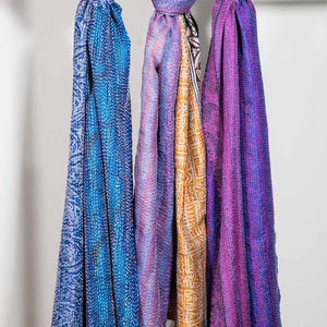 May include: Three colorful scarves hanging on a white rod. The scarves are in shades of blue, purple, and orange, with intricate patterns and fringe at the bottom. The scarves are made of a soft, flowing fabric.