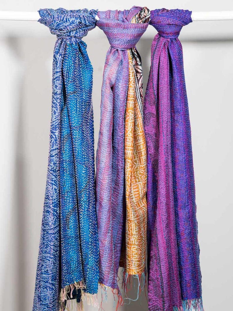 May include: Three colorful scarves hanging on a white rod. The scarves are in shades of blue, purple, and orange, with intricate patterns and fringe at the bottom. The scarves are made of a soft, flowing fabric.