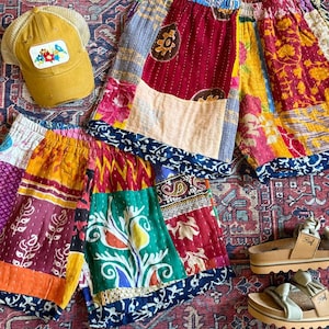Op de afbeelding: Een verzameling kleurrijke patchwork shorts met bloemen- en geometrische patronen. Een mosterdgele trucker cap met een bloemenpatch en een paar bruine sandalen zijn ook zichtbaar. De items liggen op een patroon tapijt.