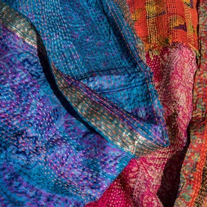May include: Close-up of a patchwork textile with vibrant colors. The fabric features intricate stitching and a mix of blue, purple, pink, orange, and yellow hues. The design includes floral and geometric patterns, creating a textured and visually rich surface.
