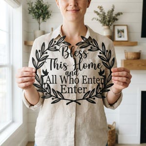 Pode incluir: Uma placa de parede circular preta com uma borda de coroa de folhas, dois pássaros pequenos e o texto Bless This Home and All Who Enter. A placa é feita de um material plano com um design recortado e letras pretas.