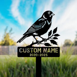 May include: Black metal bird garden stake. The bird is perched on a branch with leaves. The stake has a rectangular sign with the text "CUSTOM NAME" and the years "2020-2025". The background is a blurred view of a yard and sky.