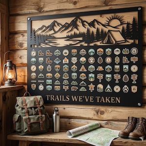 Könnte beinhalten: Ein rechteckiges Wanddisplay aus schwarzem Metall mit einer lasergeschnittenen Berg-, Wald- und Zeltlandschaft im oberen Bereich. Darunter befinden sich zahlreiche kleine, bunte Aufnäher zum Thema Outdoor. Der Text "TRAILS WE'VE TAKEN" ist in Weiß aufgedruckt.