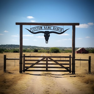 Pode incluir: Um portão de rancho de metal preto com o texto "CUSTOM NAME RANCH" acima de uma silhueta de crânio de longhorn. O portão é feito de madeira e leva a uma estrada de terra. Um celeiro vermelho é visível ao longe sob um céu azul.