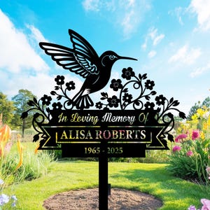 Puede incluir: Estaca de jardín conmemorativa de metal negro con un colibrí y un diseño floral. El texto dice "In Loving Memory Of ALISA ROBERTS 1965-2025". La estaca está en un jardín con flores y un cielo azul de fondo.