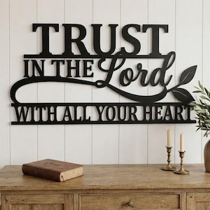 Puede incluir: Un letrero de pared de metal negro con el texto TRUST IN THE LORD WITH ALL YOUR HEART en una combinación de fuentes serif y cursiva, con un adorno decorativo y dos formas de hojas integradas en el diseño en el lado derecho.