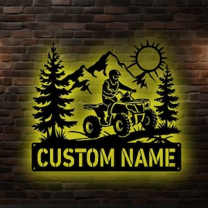 Op de afbeelding: Zwart metalen ATV-bord met een rijder, bergen, zon en bomen. Het bord heeft de tekst "CUSTOM NAME" op een rechthoekige plaat. Het bord wordt verlicht met een gele gloed tegen een bakstenen muur.