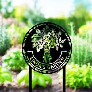 Op de afbeelding: Zwarte metalen tuinpaal met een cirkelvormig ontwerp met een boeket kruiden. De woorden "LINDA'S GARDEN" zijn in een boog rond de onderkant van de cirkel geplaatst. De kruiden zijn in groentinten en de achtergrond is een vage tuinscène.