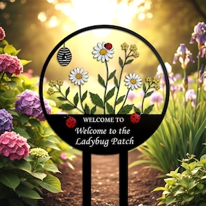 Op de afbeelding: Een zwarte metalen tuinpaal met een cirkelvormig ontwerp met madeliefjes, lieveheersbeestjes en een bijenkorf. De tekst "Welcome to the Ladybug Patch" staat onderaan. De paal is ontworpen om in een tuin te worden geplaatst.