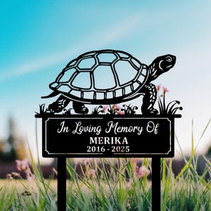 Puede incluir: Una estaca de jardín conmemorativa de metal negro con una silueta de tortuga sobre una placa rectangular. La placa dice "In Loving Memory Of MERIKA 2016-2025" en escritura blanca. La estaca está en la hierba.