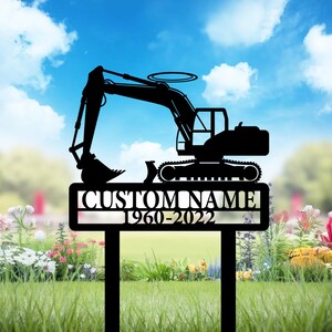 May include: Black metal excavator garden stake with the text "CUSTOM NAME 1960-2022". The excavator is a silhouette with a detailed design. The stake is in a garden with flowers and grass.