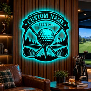 May include: A circular black metal wall sign featuring a golf ball on a tee, crossed golf clubs, and a forest landscape silhouette. The sign is illuminated with a bright cyan LED backlight and displays the text CUSTOM NAME and TEE TIME.