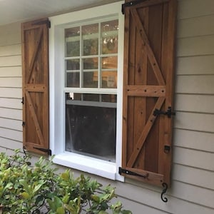May include: Exterior view of a window with wooden shutters. The shutters are stained a warm brown and feature a diagonal brace design. Black metal hinges and hardware are visible. The window frame is white, and the house siding is a light beige.
