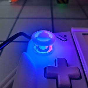 May include: Close-up of a white handheld gaming console with a glowing blue joystick and a purple directional pad. The joystick emits a bright blue light, with a pink ring around its base. A black cable is visible.