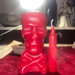 May include: A red skull-shaped candle with a lit wick, next to a smaller red candle. The skull candle has a detailed face and a crossbones design. The candles are on a red velvet surface.