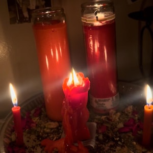 Könnte beinhalten: Mehrere brennende rote Kerzen unterschiedlicher Größe, darunter Säulen- und Spitzkerzen, auf einem silbernen Tablett. Die Kerzen sind von getrockneten Kräutern und Blütenblättern umgeben. Im Hintergrund befindet sich ein gerahmtes Schwarz-Weiß-Foto.