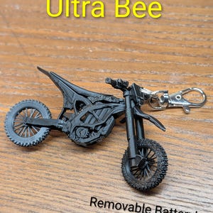 Op de afbeelding: Zwarte Ultra Bee motorfiets sleutelhanger met een uitneembaar batterijontwerp. De sleutelhanger is gemaakt van een donker materiaal en heeft gedetailleerde wielen en een zilveren sluiting. Tekst op de afbeelding luidt "Ultra Bee" en "Removable Battery!"