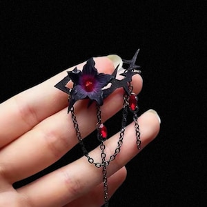 Puede incluir: Un accesorio para el cabello negro con una flor morada oscura, detalles de gemas rojas y cadenas colgantes. El diseño incluye formas geométricas y se sostiene en una mano sobre un fondo negro. El texto "Shoppshoow" es visible en la parte inferior.