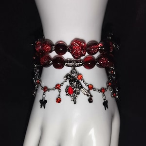 Puede incluir: Una pulsera de dos hilos con cuentas rojo oscuro y un dije decorativo. La hebra superior tiene cuentas grandes y redondas y una cuenta en forma de rosa. La hebra inferior tiene un dije colgante con detalles rojos y pequeños dijes negros de mariposa y diente.