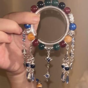 Puede incluir: Se muestra una pulsera de cuentas multicolores y un brazalete a juego. Las pulseras presentan una variedad de cuentas en colores como rojo, azul, amarillo, verde y negro. Los dijes plateados y los acentos colgantes añaden detalles.