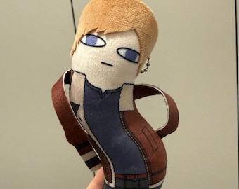 May include: A plush doll featuring a character with short light brown hair, blue eyes, and a neutral expression. The doll wears a dark blue shirt, a brown jacket with white trim, and dark blue pants. A small metal ball chain is attached to the head.