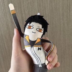 May include: A plush doll featuring a character with black hair, orange eyes, and a simple embroidered face. The doll wears a white and dark grey outfit with yellow accents and a black letter N on the chest. It has a long, curved body shape.