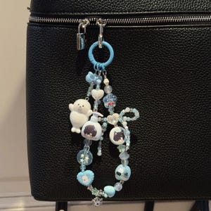 May include: A light blue beaded keychain with various charms, including a white plush seal, heart-shaped beads, and round beads with cartoon images. The keychain is attached to a black bag.