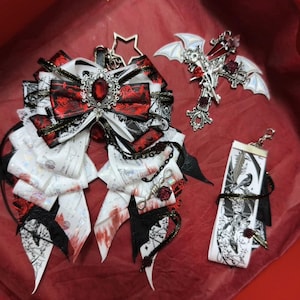 Puede incluir: Conjunto de accesorios de temática gótica, que incluye un lazo grande con detalles en rojo y negro, un colgante de cruz con alas de murciélago y un llavero de cinta. El lazo presenta una joya central y detalles intrincados.