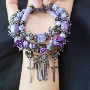 Puede incluir: Una pulsera de cuentas moradas con elementos decorativos. La pulsera presenta varios tonos de cuentas moradas, detalles de mariposas y colgantes de espadas. El diseño incluye detalles florales y ornamentales, creando una estética gótica o fantástica.
