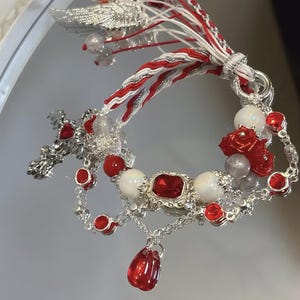 May include: A silver-toned bracelet with red and white accents, featuring red gemstones, white beads, and a dangling red teardrop pendant. The bracelet has a cross and wing design, with a braided cord and the text "Shoppshoow".
