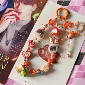 Puede incluir: Una pulsera de abalorios con varios dijes, incluyendo corazones, manzanas e ilustraciones de personajes. La pulsera presenta cuentas naranjas, rojas y transparentes, con un cierre de mosquetón. El fondo incluye una postal con una ilustración masculina y la palabra "Forever".
