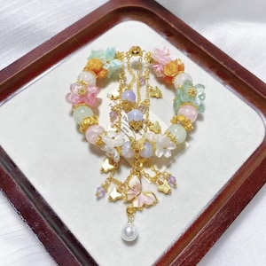 Puede incluir: Una pulsera de colores pastel con abalorios de flores y mariposas. La pulsera presenta una mezcla de cuentas rosas, verdes y amarillas, con detalles dorados. Cuelgan de la pulsera abalorios de mariposas y un colgante de perlas.