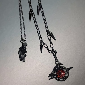 Puede incluir: Collares colgantes de metal negro. Uno presenta un dije de pluma y una pequeña cuenta. El otro tiene una cadena con púas y un colgante con una gema roja y un diseño de pájaro. La marca "Shoppshoow" está en la parte inferior.