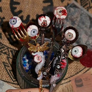 Puede incluir: Una colección de cucharas y tenedores decorativos, cada uno adornado con un diseño de ojo realista. Los utensilios se exhiben en un tarro de cristal lleno de cuentas azules y transparentes. Los mangos presentan detalles y adornos ornamentados.