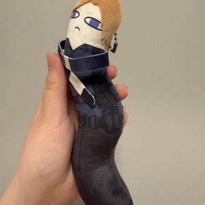 May include: A soft plush toy shaped like a long, curved cylinder featuring a character with light brown hair and blue eyes. The body is printed with a dark blue tactical vest, black utility pouches, and dark gray pants. A metal ball chain is attached.
