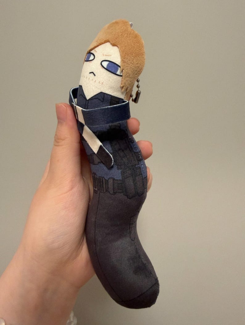 May include: A soft plush toy shaped like a long, curved cylinder featuring a character with light brown hair and blue eyes. The body is printed with a dark blue tactical vest, black utility pouches, and dark gray pants. A metal ball chain is attached.