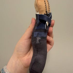 May include: A plush character keychain featuring a tan head and a dark blue and charcoal gray outfit with printed belt and axe details. The soft figure includes a silver metal ball chain attached to the top for hanging, showcasing a detailed design.