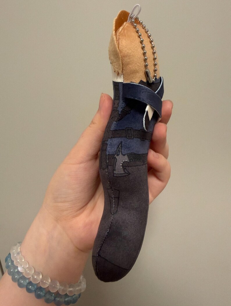 May include: A plush character keychain featuring a tan head and a dark blue and charcoal gray outfit with printed belt and axe details. The soft figure includes a silver metal ball chain attached to the top for hanging, showcasing a detailed design.