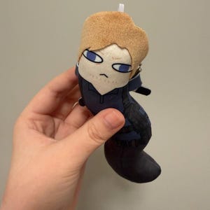 May include: A small plush doll featuring a character with light brown hair, blue eyes, and a serious expression. The doll wears a dark blue outfit with black tactical-style straps and a small white loop at the top of the head for hanging purposes.