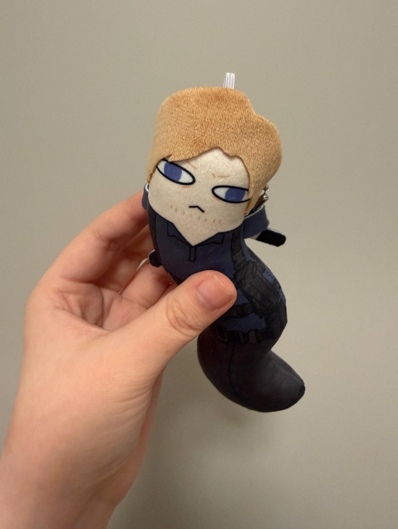 May include: A small plush doll featuring a character with light brown hair, blue eyes, and a serious expression. The doll wears a dark blue outfit with black tactical-style straps and a small white loop at the top of the head for hanging purposes.