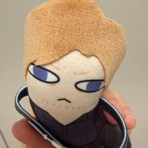 May include: A plush toy featuring a character with short tan hair, light skin, and purple eyes. The figure has a small mouth, light brown stubble, and wears a dark blue collared shirt. A small white loop is attached to the top of the head.