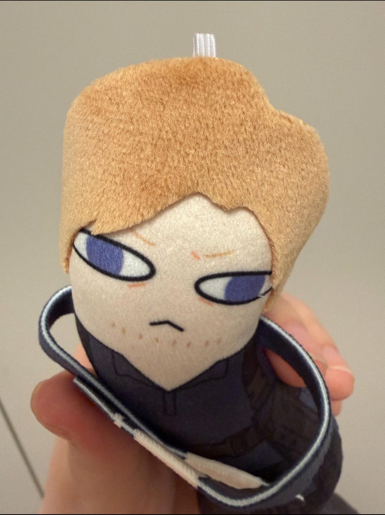 May include: A plush toy featuring a character with short tan hair, light skin, and purple eyes. The figure has a small mouth, light brown stubble, and wears a dark blue collared shirt. A small white loop is attached to the top of the head.