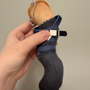 May include: A plush character keychain featuring a tan head with orange hair, a dark blue shirt with a black belt, and dark gray pants. The figure includes a silver metal ball chain attached to the top for hanging, with a printed fabric design.